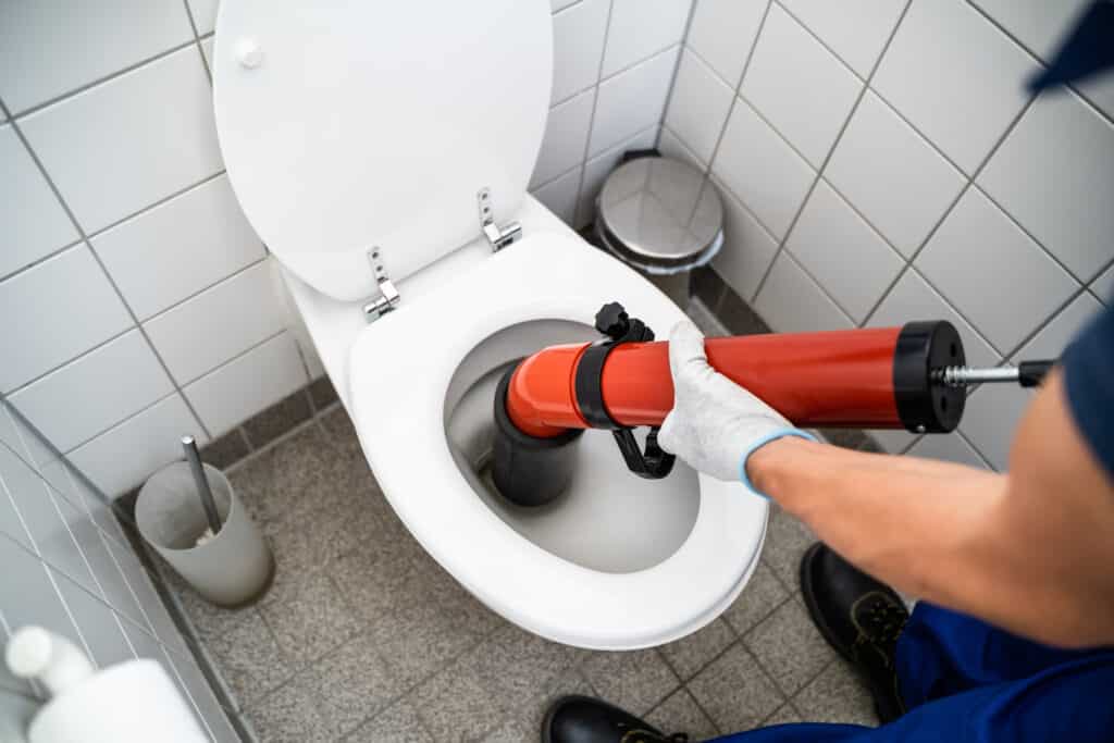 Débouchage toilettes : Plombier professionnel intervient Plombier avec gants blancs utilise un déboucheur professionnel rouge et noir pour déboucher des toilettes blanches. Carrelage mural blanc et sol gris.