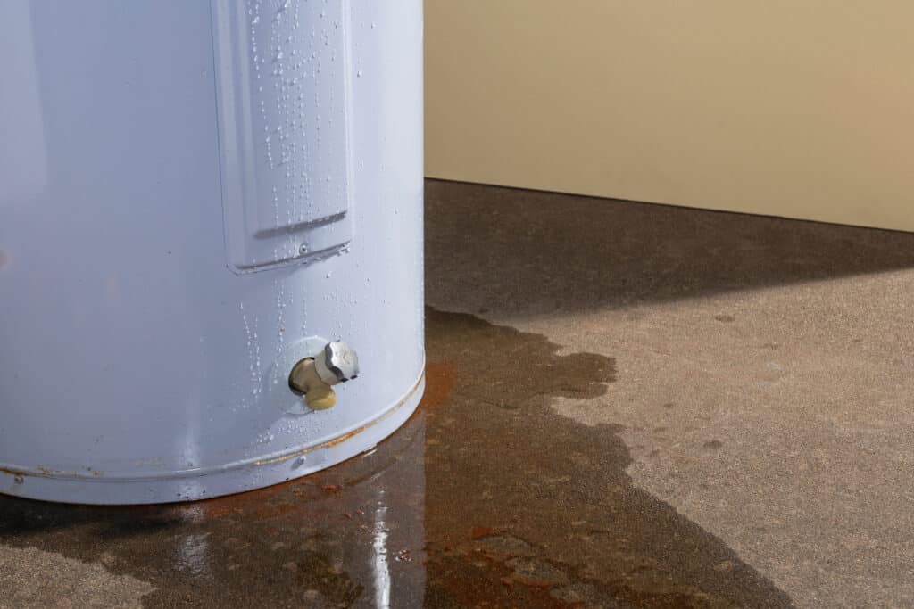Chauffe-eau blanc fuyant avec gouttes d'eau ruisselantes et flaque rouillée sur sol en béton.