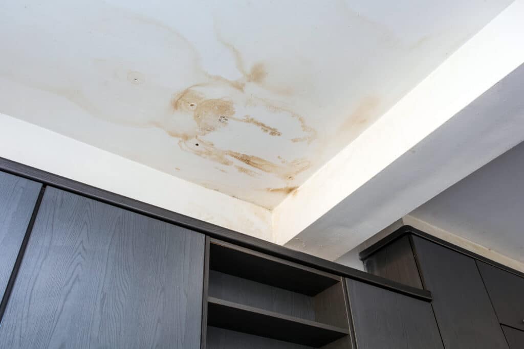 Plafond blanc avec de larges taches d'eau marron suite à une fuite, surmontant des meubles de rangement sombres. Indique des dégâts.