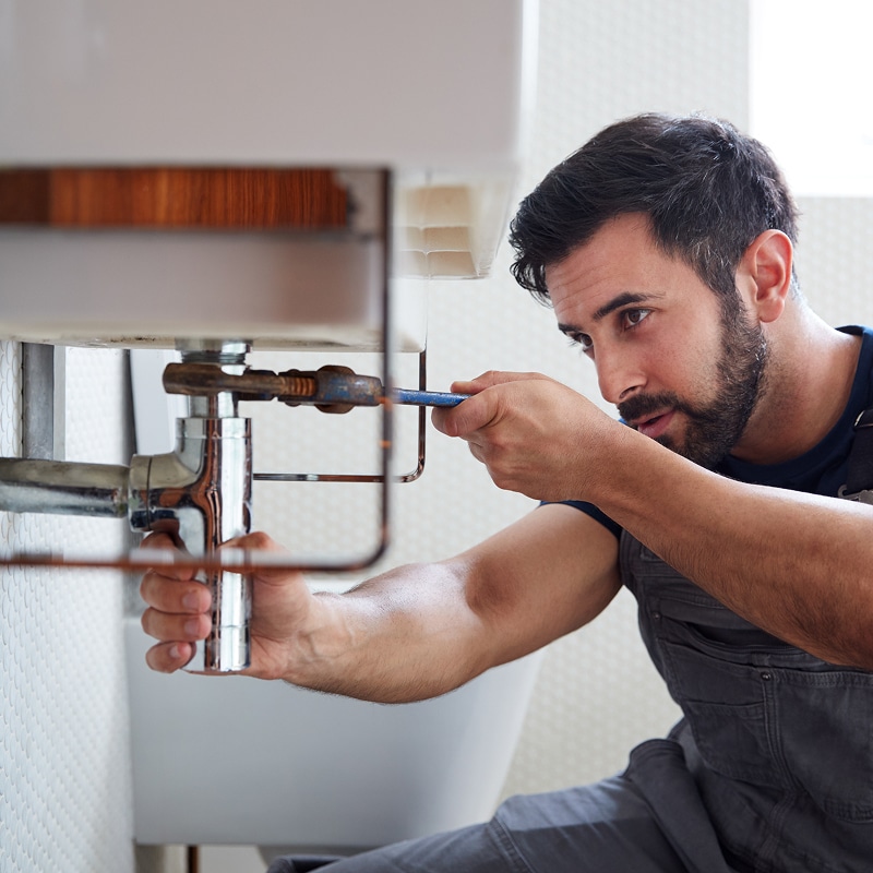 Plombier barbu concentré réparant un tuyau sous un évier de salle de bain avec une clé. Travail de maintenance qualifié.