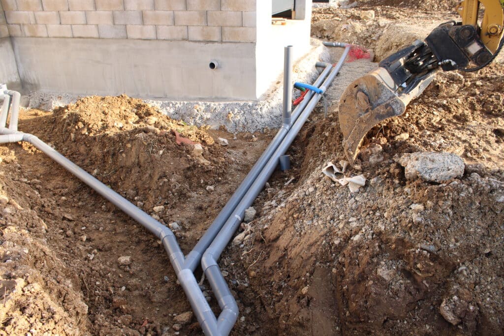 Fondation de bâtiment avec tuyaux PVC gris posés dans des tranchées. Chantier d'excavation en cours pour raccordements.
