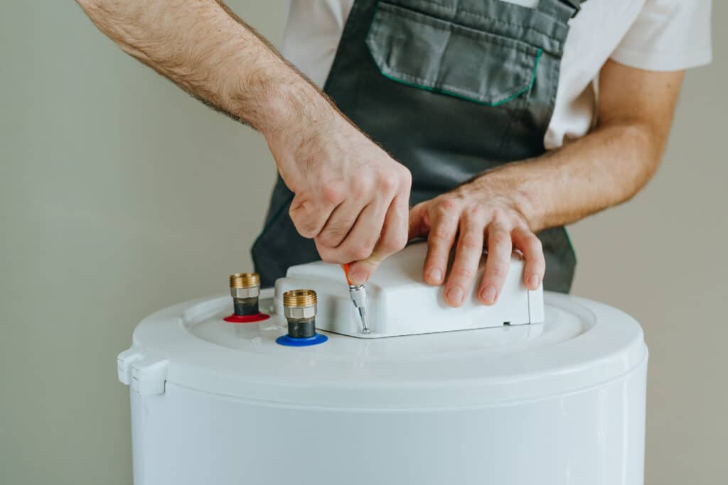 Plombier réparant un chauffe-eau blanc avec un tournevis, ses mains ajustant un composant près des raccords d'eau.