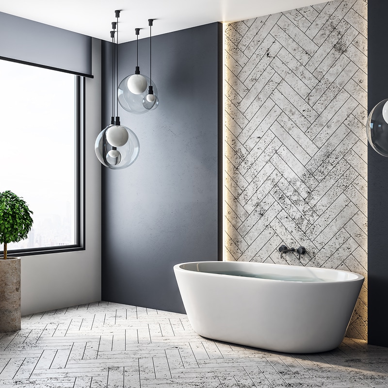 Salle de bain moderne grise, baignoire blanche, carrelage chevrons clair, suspensions sphériques et grande fenêtre urbaine.