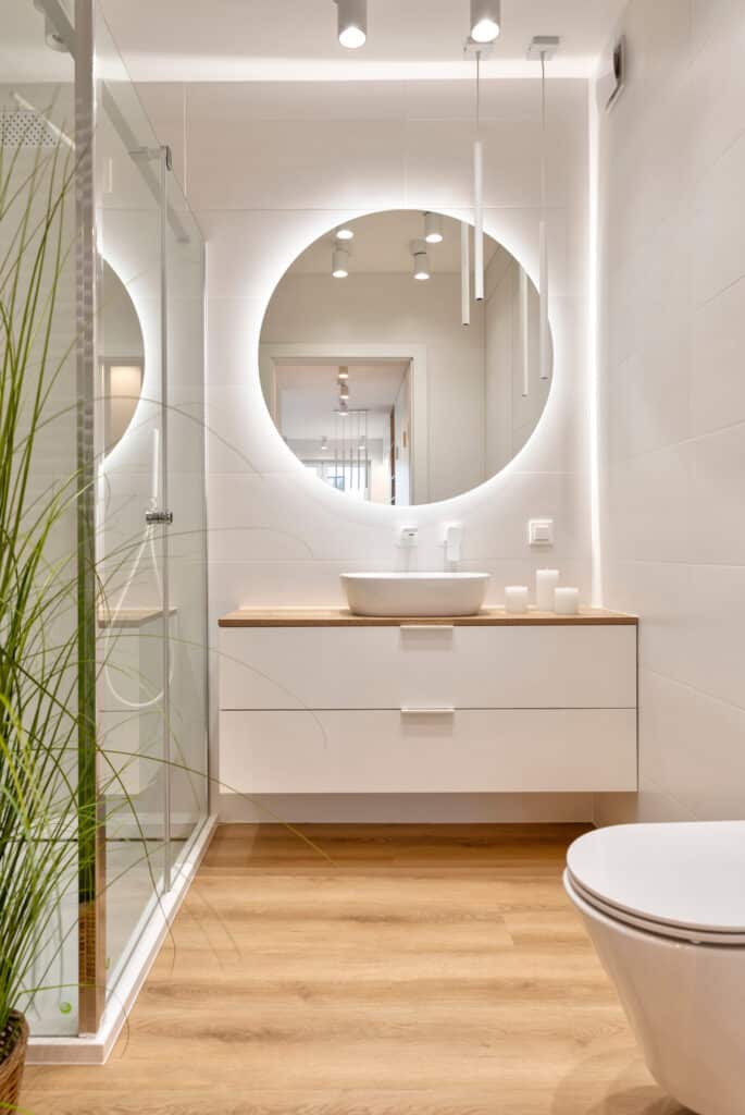 Intérieur de salle de bain moderne épurée en blanc et bois clair, avec douche en verre, meuble-lavabo suspendu et grand miroir rond rétroéclairé.