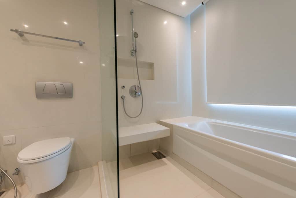 Salle de bain moderne épurée avec douche en verre, toilettes suspendues et baignoire blanche, carrelage clair, éclairage LED.